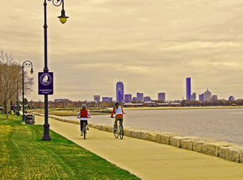 Bikers along Dorchester Bay. The Boston Harborwalk sign-board in the forefront with the Boston skyline in the distance.