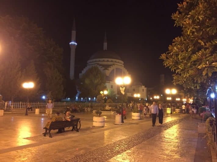 Pedestrians on Kolë Idromeno Street in Shkodër at night, with Ebu Bekr Mosque in the background