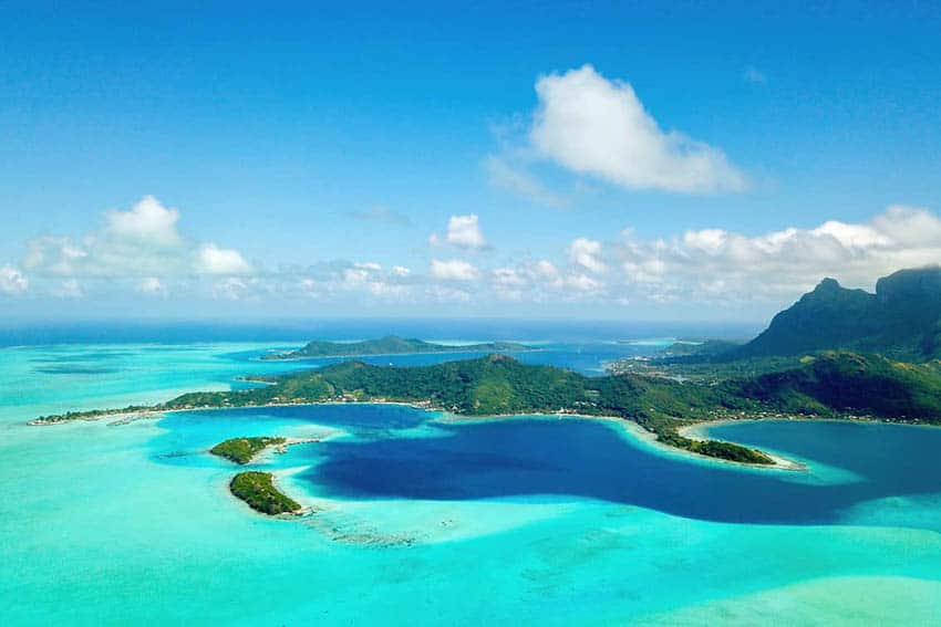 Bora Bora Island creeps into the shot and casts a shadow over the sea, as white clouds fade into the background.