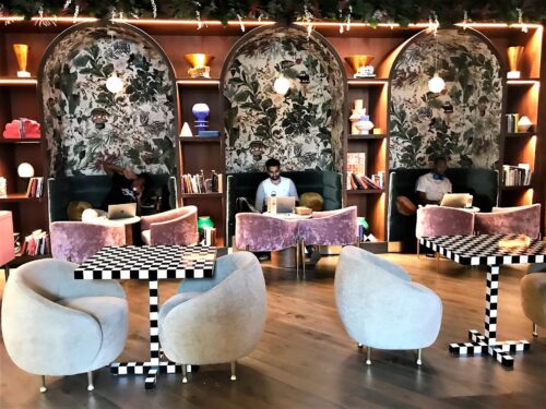 Coved banquette seating lined in wowy wallpaper, plush velvet chairs, and checkerboard tables make the Funny Library & Coffee Bar a favorite spot to hang out and work or have coffee with friends at Virgin Hotel Dallas.