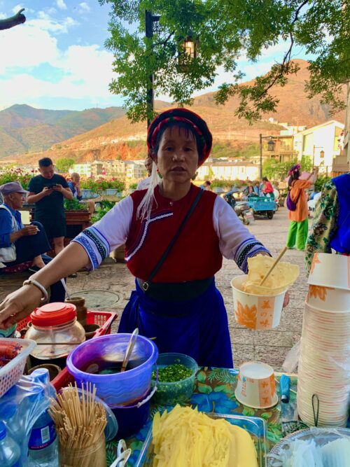 Woman in Bai ethnic clothing sells fried milk fans, topped with sweet or savory sauce.